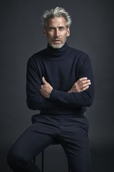 an older man with grey hair sitting on a stool in front of a black background