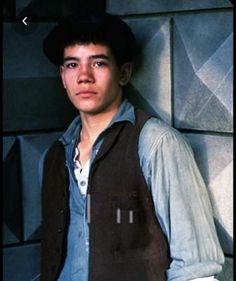 a young man leaning against a wall wearing a black hat and vest with his hands in his pockets