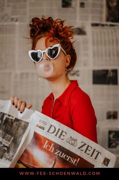 a woman wearing sunglasses and blowing bubbles while holding a newspaper in front of her face