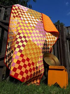 a colorful quilt sitting on the ground next to a yellow trash can and wooden fence