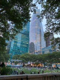 people are sitting on the grass in front of skyscrapers and trees at dusk time