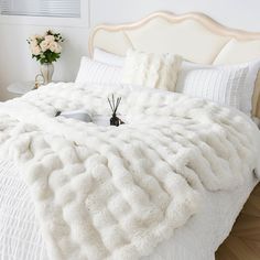 a large white bed covered in blankets and pillows with flowers on the side table next to it