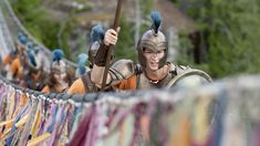 a man dressed in roman armor holding a stick and walking on a rope bridge with other people behind him