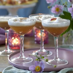 three glasses filled with drinks sitting on top of a pink platter next to flowers