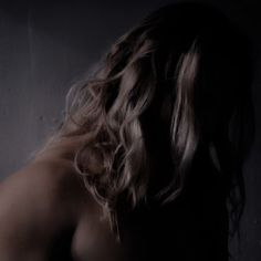 a man with long hair and no shirt on standing in front of a gray wall