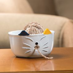 a white cat shaped bowl with balls of yarn in it on top of a wooden table