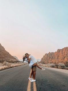 a man and woman kissing on the side of an empty road with mountains in the background