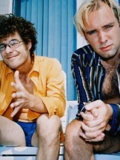 two men sitting next to each other in front of a blue wall with one pointing at the camera