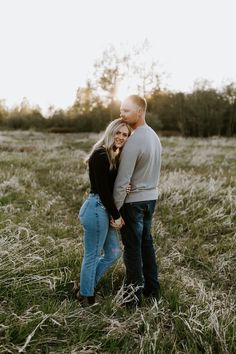 an engaged couple standing in the middle of a field at sunset with their arms around each other