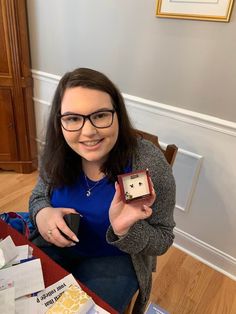 a woman sitting at a table holding up a small box with a clock on it