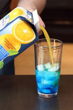 a person pouring blue liquid into a glass with an orange slice on top and ice in the bottom