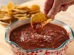 a hand dipping a tortilla chip into a bowl of salsa with chips on the side