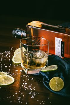 a glass filled with ice and limes next to a bottle of whiskey on a wooden table