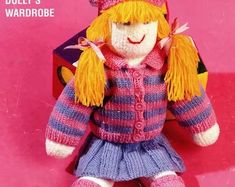 a knitted doll with blonde hair wearing a pink and blue sweater, purple pants and hat