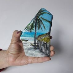 a hand holding a rock with a painting of a beach and palm trees on it