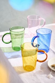 three different colored glass mugs sitting on top of a white cloth covered tablecloth