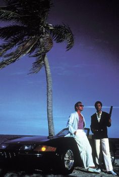 two men standing next to a car on the beach with a palm tree in the background