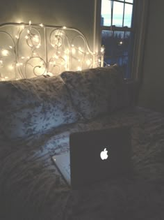 an apple laptop sitting on top of a bed next to a window with fairy lights