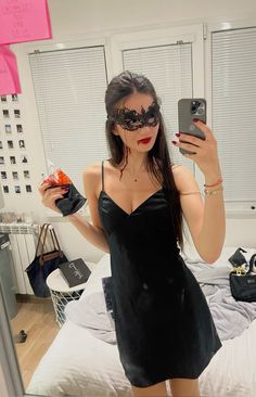 a woman in a black dress taking a selfie with her cell phone and wearing a masquerade