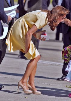 a woman in a yellow dress bending over to pick something out of the ground with her hand