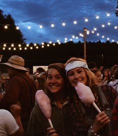 two young women are holding cotton candy at an outdoor event with string lights in the background