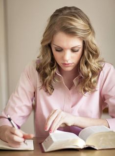 a woman sitting at a table with an open book and pen in her hand as she writes