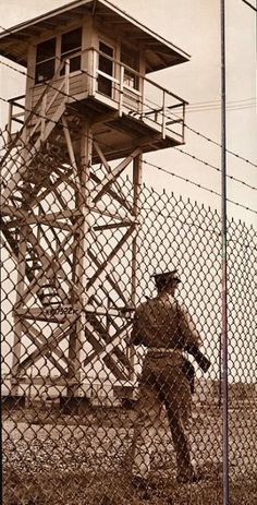 an old photo of a man standing in front of a guard tower behind a chain link fence