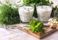 two glasses filled with yogurt sitting on top of a cutting board next to fresh herbs