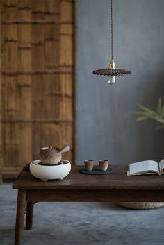 a wooden table topped with two cups and a book next to a light hanging from the ceiling