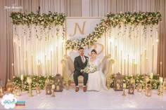 a bride and groom sitting on a chair in front of a wedding backdrop with candles