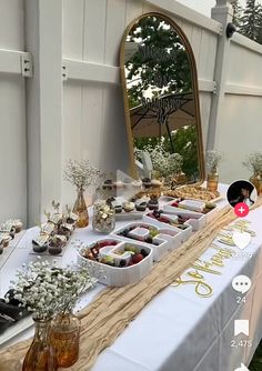 a table topped with lots of food next to a mirror and trees in the background