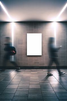 two people walking past a white square sign on a wall in an empty room with tile flooring