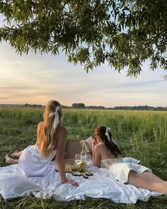 two women in white dresses sitting on a blanket under a tree and having drinks together