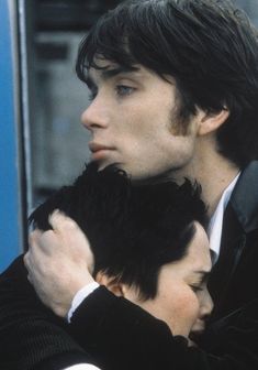 two people hugging each other in front of a blue wall and one person wearing a suit