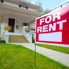 a red for rent sign sitting in front of a white house with grass on the lawn