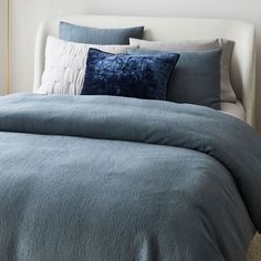 a bed with blue comforter and pillows in a white walled room next to a window