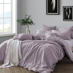 a bed with purple sheets and pillows in a room next to a white chair, potted plant and framed pictures on the wall