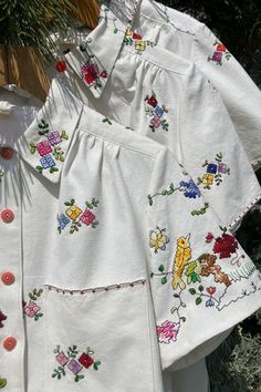 two white blouses with colorful flowers on them hanging from a clothes line in front of some trees