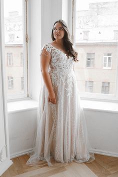 a woman standing in front of a window wearing a wedding dress and posing for the camera