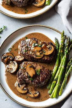 two white plates topped with steak, mushrooms and asparagus covered in gravy