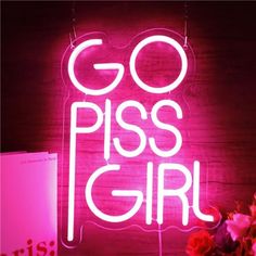 a pink neon sign that says go piss girl with flowers in front of it on a table