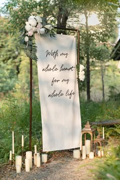 a wedding sign with candles and flowers on the ground in front of it, surrounded by greenery