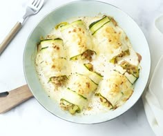 a dish with zucchini and cheese in it on a white table next to a wooden utensil