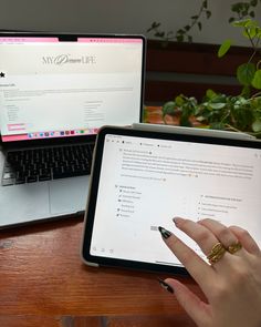 a person using a tablet computer on a wooden table with a laptop and potted plant in the background