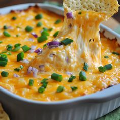 a tortilla chip being dipped with cheesy cheese and green onions in a casserole dish