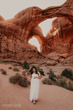 a woman in a white dress standing in front of an arch shaped rock formation at sunset