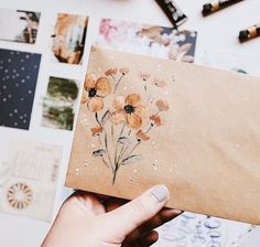 a person holding a piece of paper with flowers painted on it, surrounded by other papers and pens