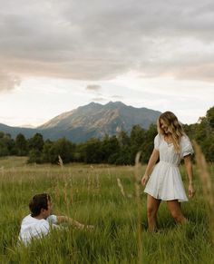 a woman in a white dress standing next to a man in a grassy field with mountains in the background