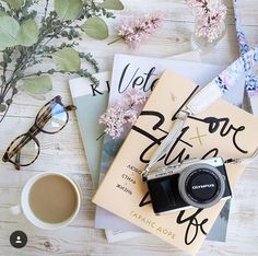 a camera, book and cup of coffee on top of a wooden table next to flowers