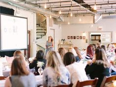 a group of people sitting at tables in front of a projector screen with a woman standing on it